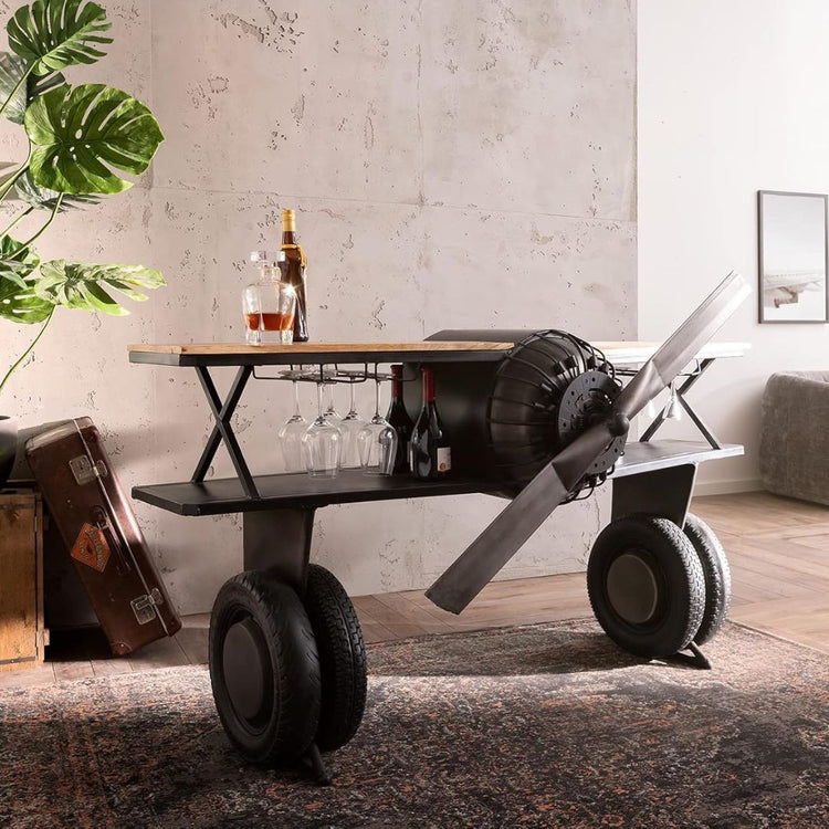Industrial airplane-engine style bar cart with propeller decoration and dual shelves, featuring wooden tabletop, black metal frame, large rubberized wheels, hanging wine-glass rack and storage for bottles, set in a modern living-room environment