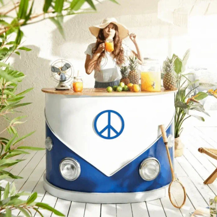 A camper van-themed bar counter with a blue and white color scheme, featuring headlights and a peace symbol emblem, set in a tropical-style setting with various plants and a pineapple.