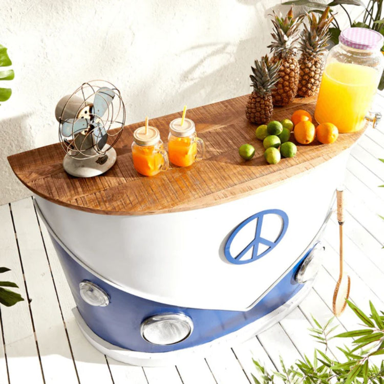 A camper van-themed bar counter with a blue and white color scheme, featuring headlights and a peace symbol emblem, set in a tropical-style setting with various plants and a pineapple.